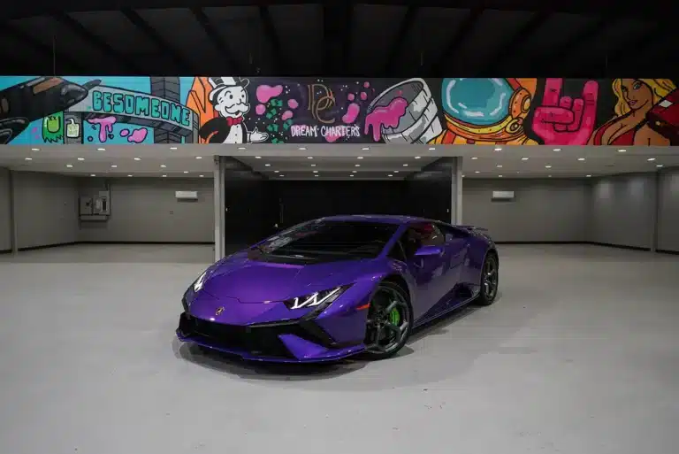 A purple Lamborghini sports car is parked in an indoor, well-lit showroom with a colorful graffiti mural above featuring various cartoon and artistic elements.