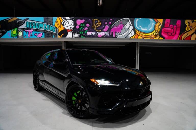 A sleek black Lamborghini SUV, available for renting a Lamborghini experience, is parked indoors under a colorful, graffiti-style mural featuring cartoon characters, bold shapes, and vibrant colors. The car’s wheels and windows reflect the overhead lighting.